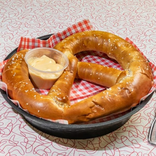 A large, salted pretzel with a side of cheese dip, served in a black pan on a retro tablecloth.