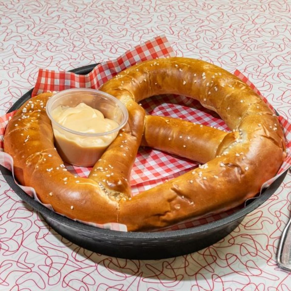 A large, salted pretzel with a side of cheese dip, served in a black pan on a retro tablecloth.